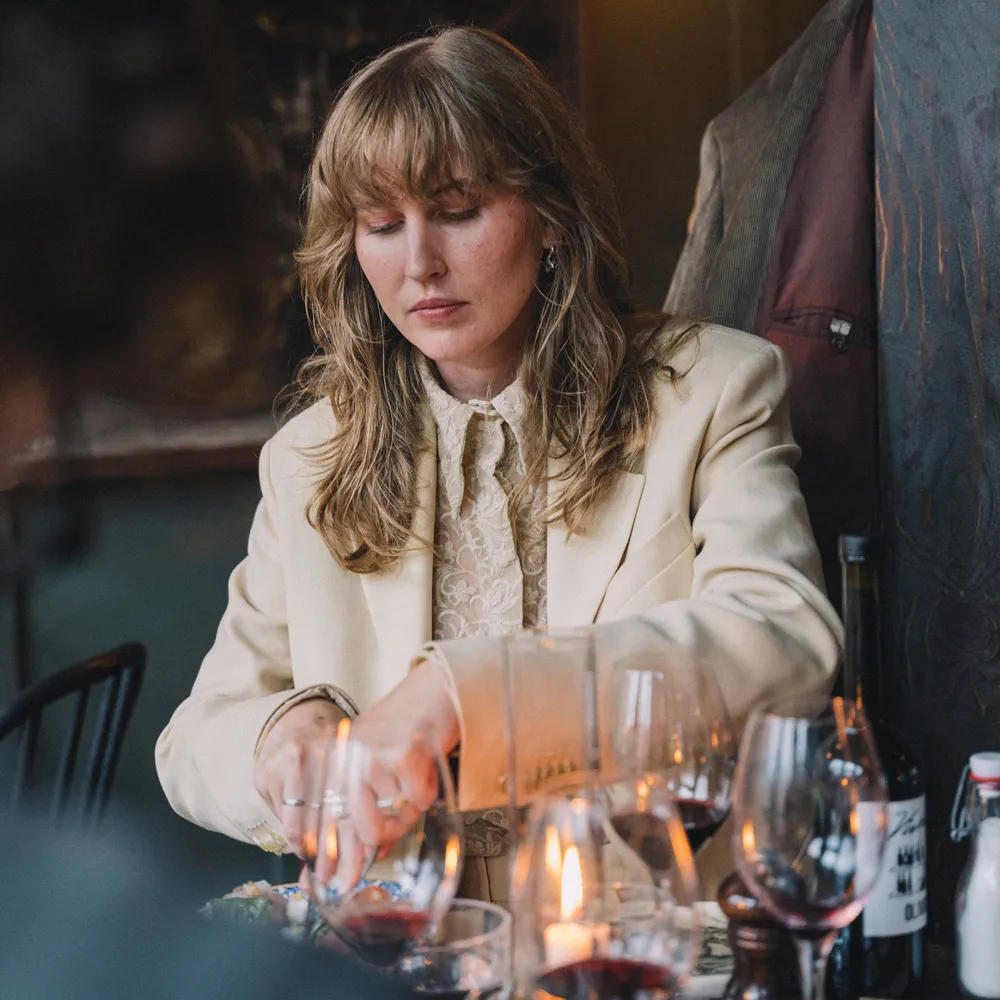 Kvinna i krämfärgad blazer och spetsblus som äter vid ett stearinljusbelyst bord med glas och flaskor med rött vin.
