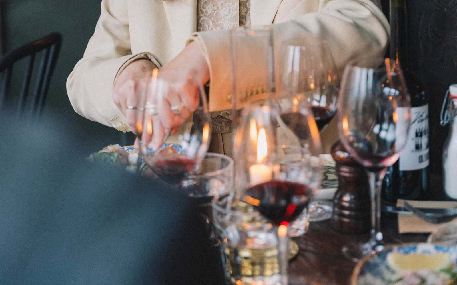 Person med beige jacka och ringar på fingrarna sitter vid ett dukat bord med tända ljus, glas med rött vin och en vinflaska.