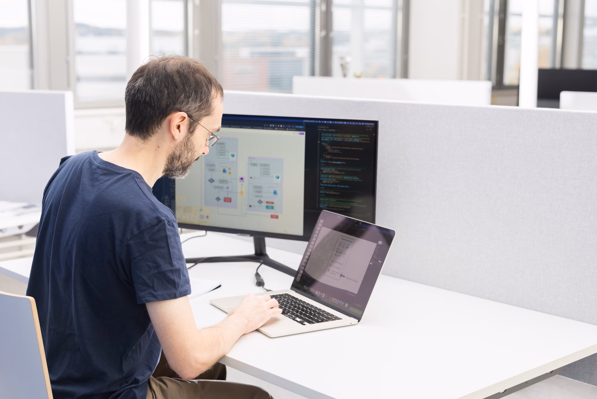 Man with glasses and a beard working on a laptop at a white office desk with an additional monitor displaying diagrams and code, in a bright modern office environment.