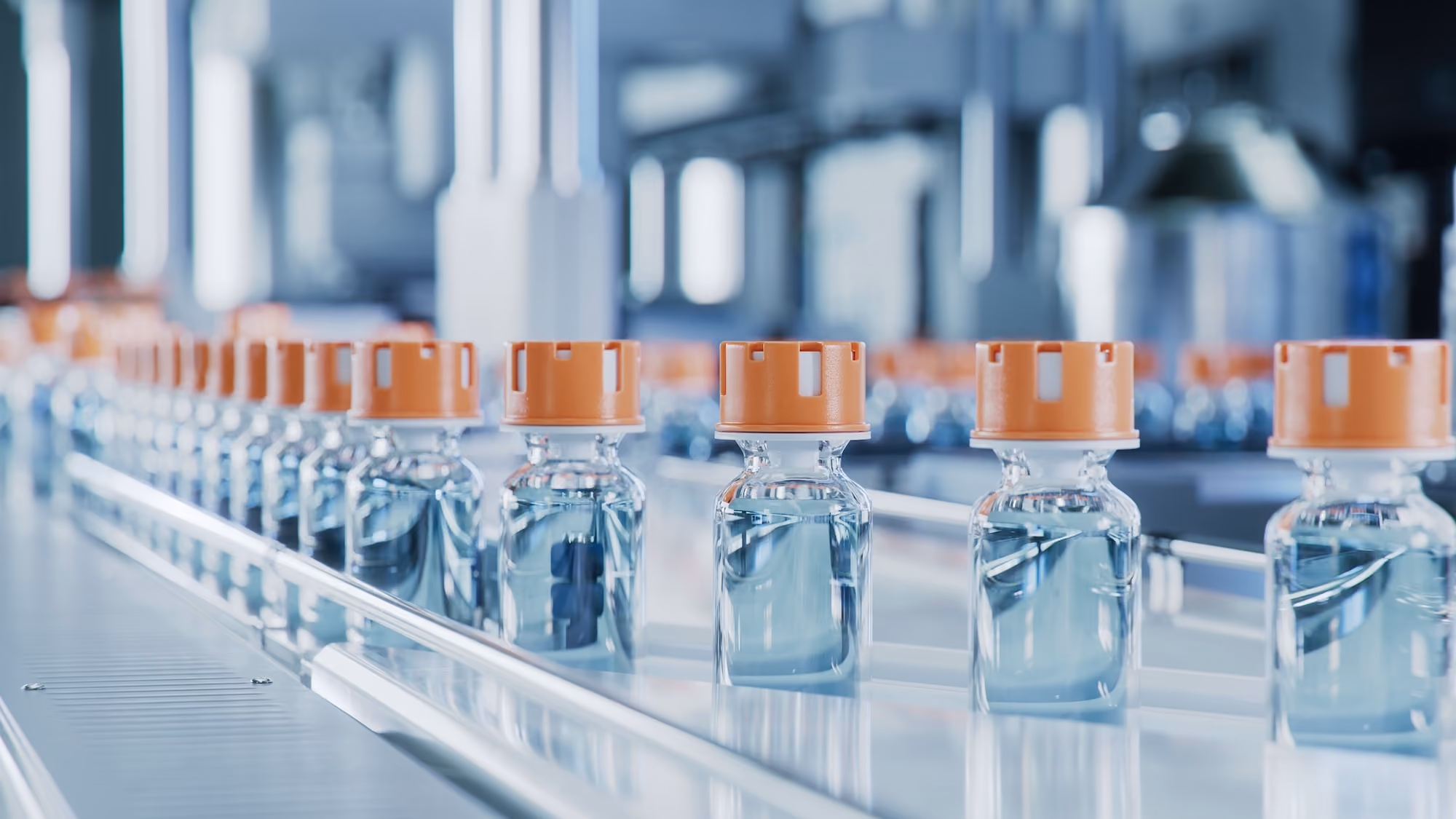 A row of small glass vials filled with clear liquid and sealed with orange caps moving on a conveyor belt in a pharmaceutical production facility.