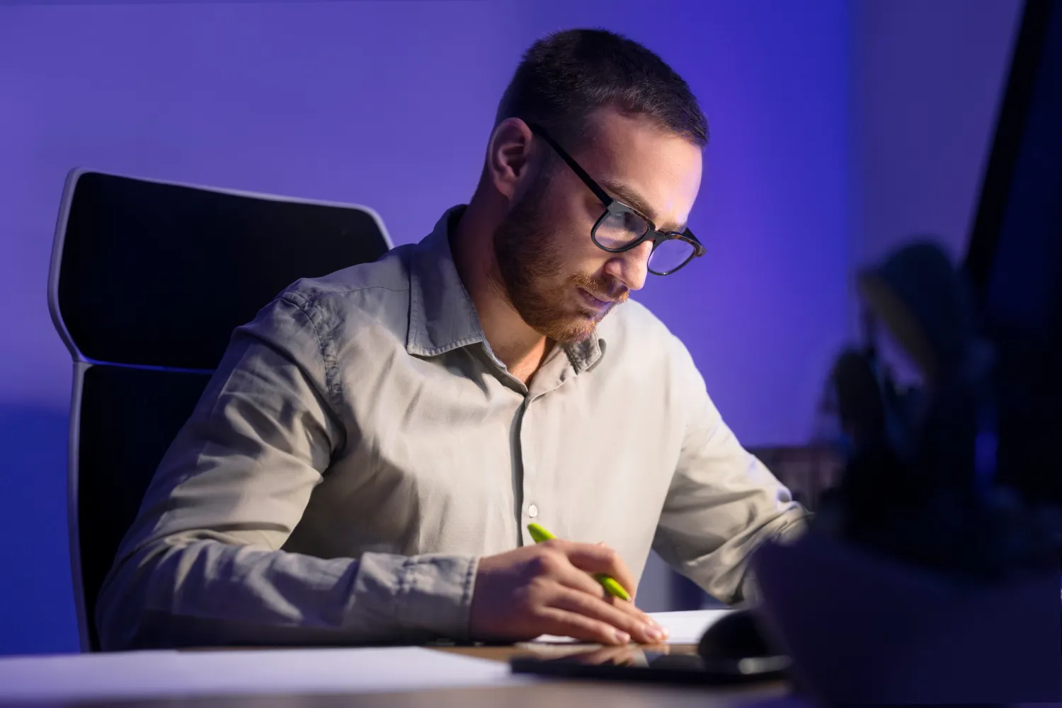 Man with glasses writing on paper with a green pen while sitting in a dark room with blue lighting.