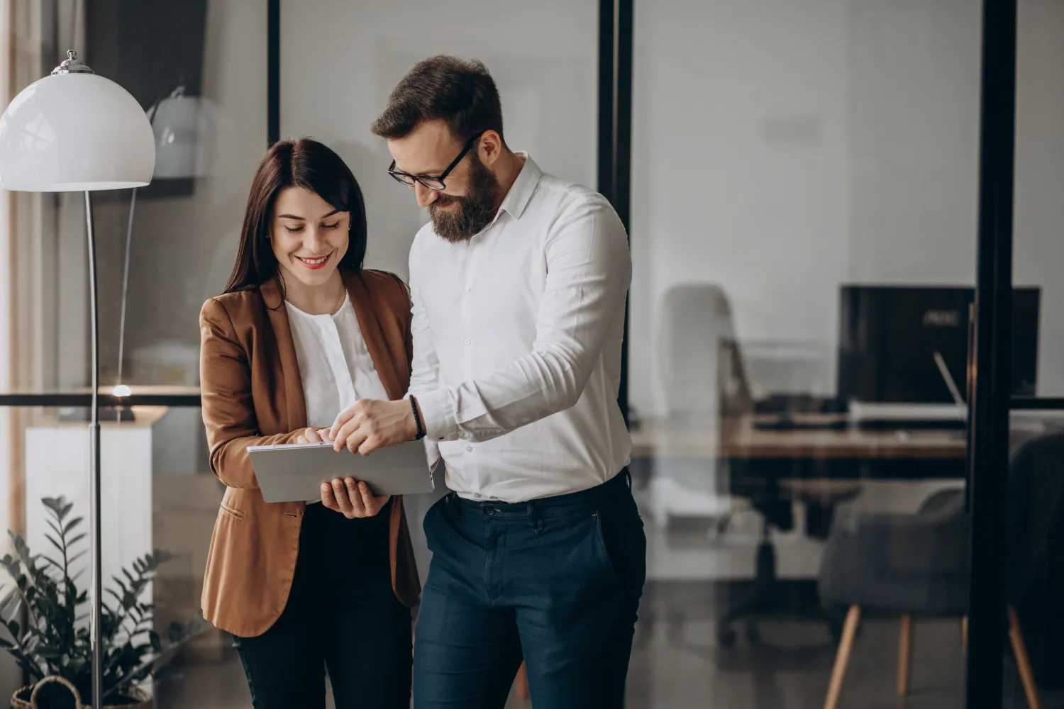 A man and a woman standing in an office, smiling and looking at a tablet together.