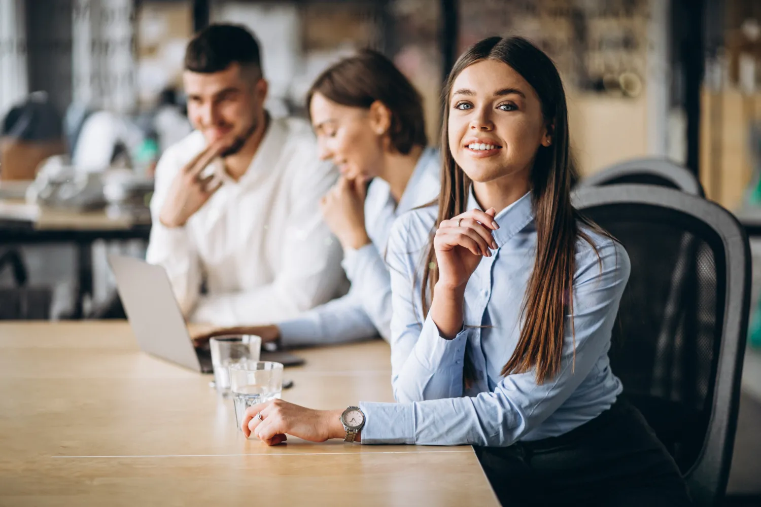 Smiling woman in blue shirt sitting at a desk with two colleagues working on a laptop in the background.