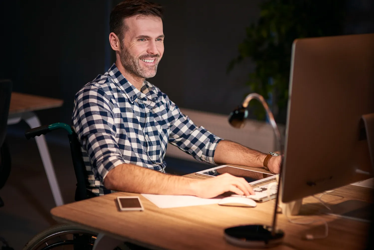 Smiling man in a wheelchair working on a desktop computer at a desk with a smartphone and tablet nearby.
