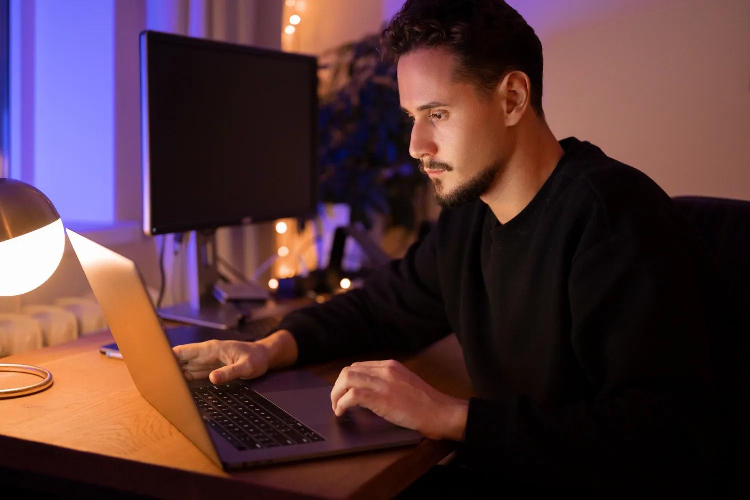 Man focused on working on a laptop at a desk illuminated by a warm desk lamp in a dimly lit room.