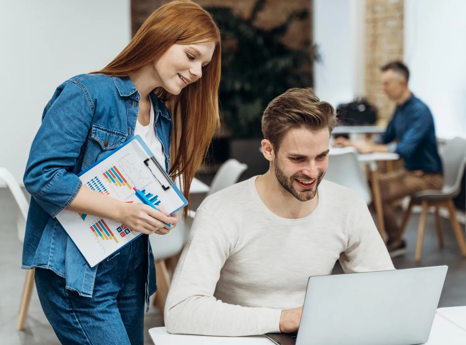 A woman holding charts on a clipboard is standing next to a man who is smiling and working on a laptop in an office setting.
