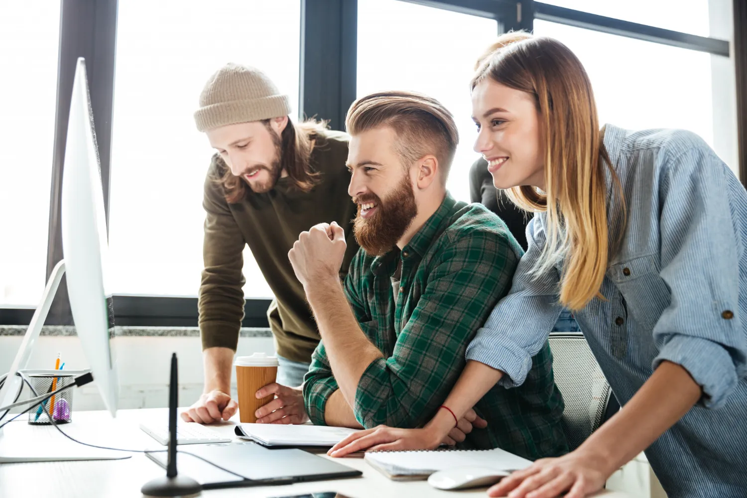 Three young coworkers smiling and looking at a computer screen in a bright office.