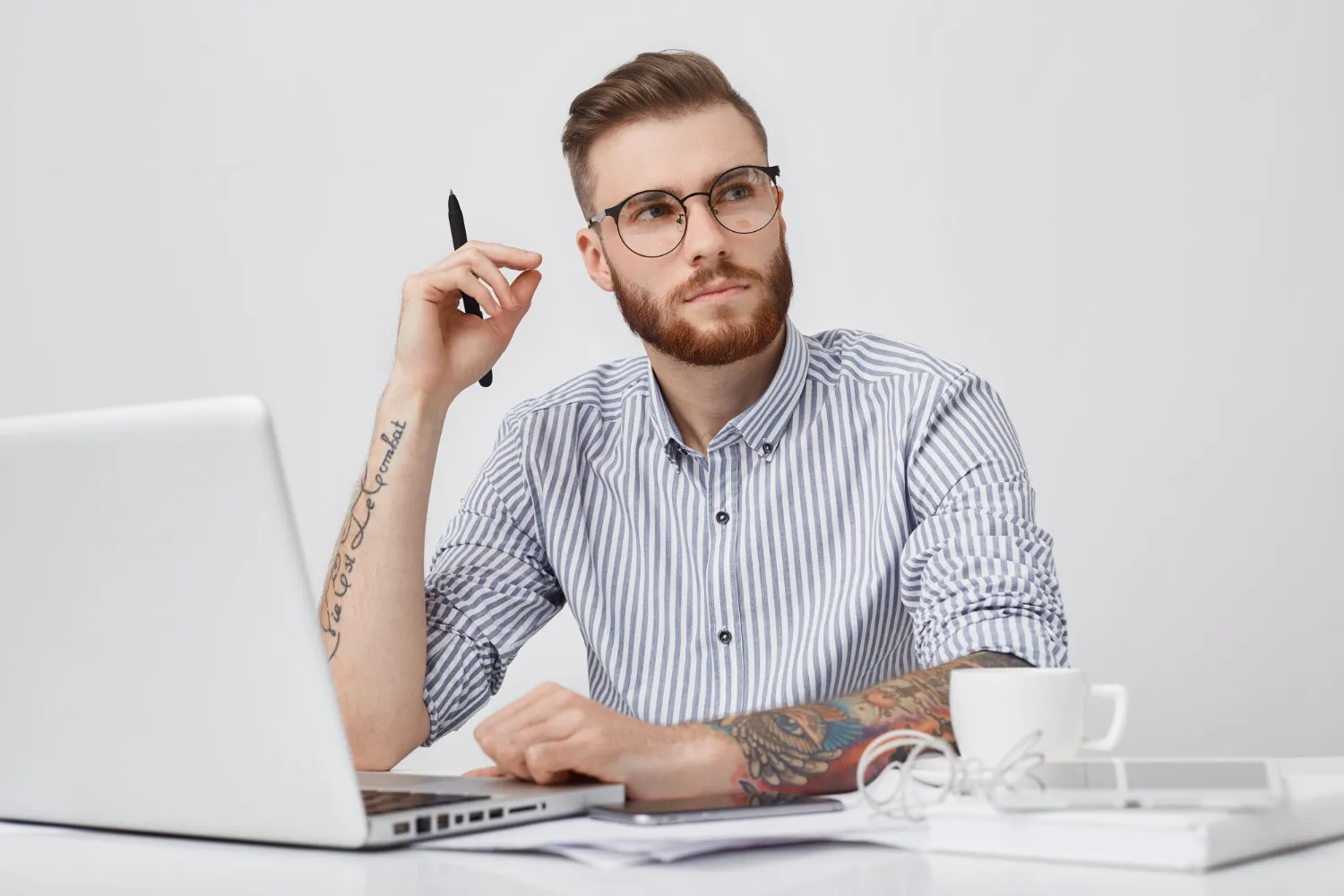 Bearded man with glasses and tattoos holding a pen, sitting at a desk with laptop, coffee cup, and tablet, looking thoughtfully to the side.