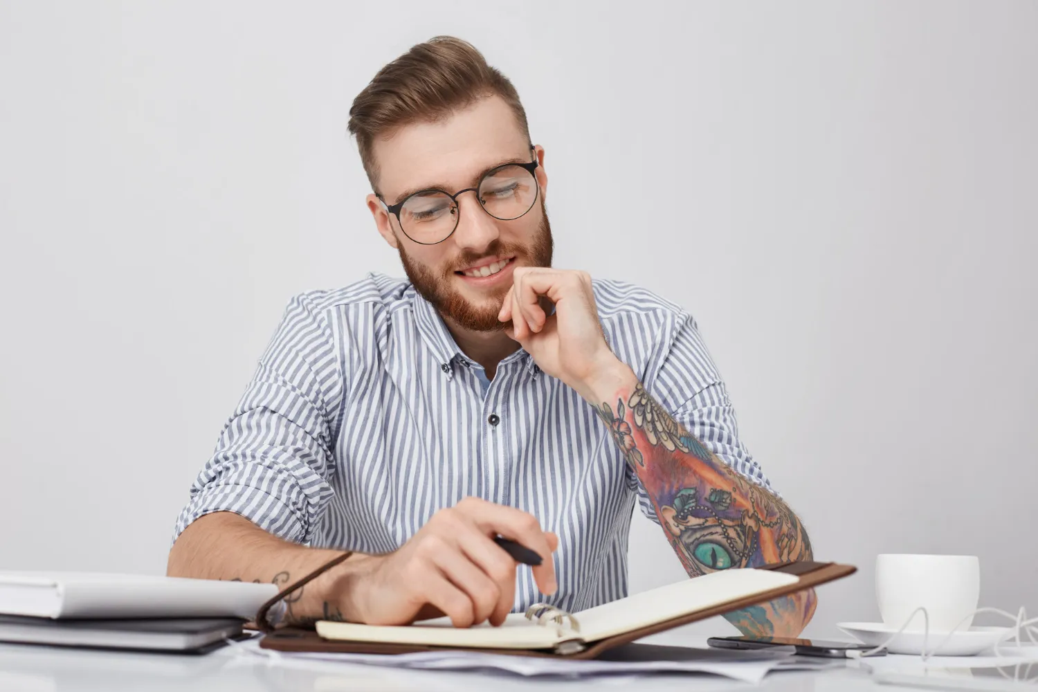 Smiling young man with glasses and a tattooed arm writing in a notebook at a desk with coffee cup and papers.