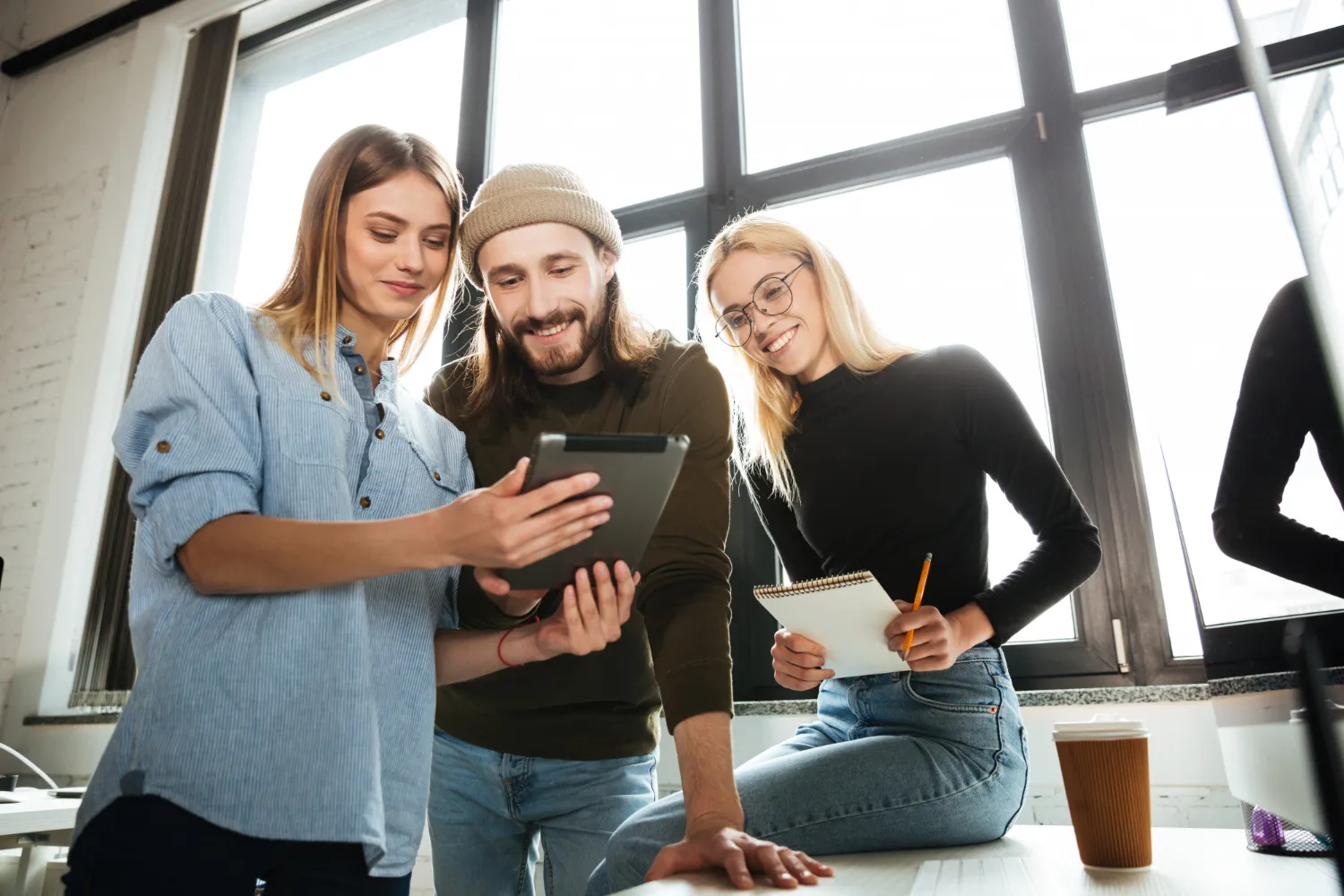 Three young people smiling and looking at a tablet in a bright office with large windows.