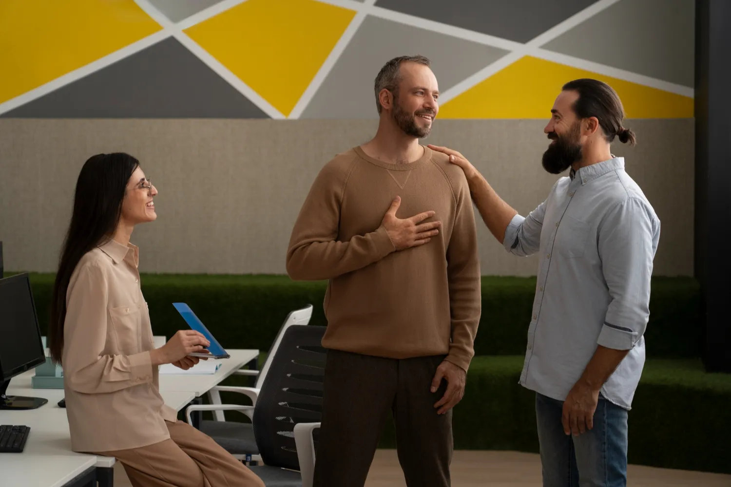 Three colleagues interact positively in an office, with one man smiling and placing a hand on his chest as another man touches his shoulder while a woman sits nearby smiling and holding a tablet.