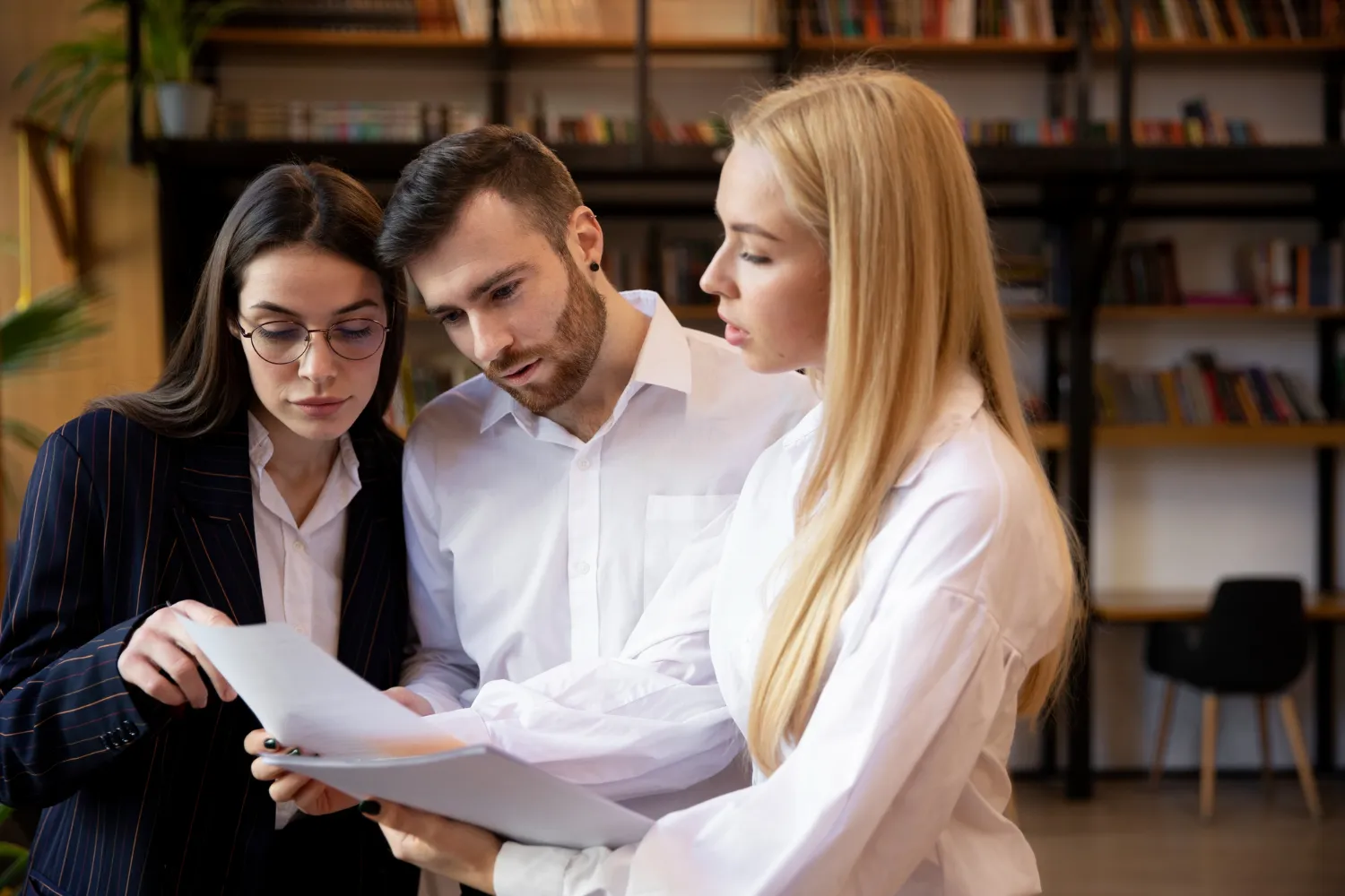 Three young professionals closely reviewing documents together in a modern office with bookshelves in the background.