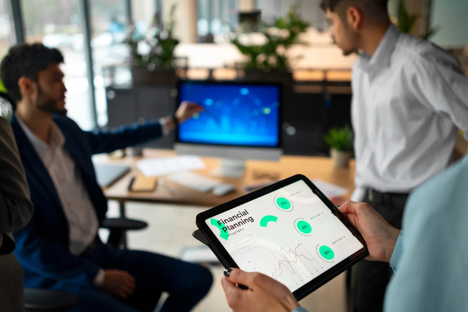 Person holding a tablet displaying financial planning charts with graphs and percentages, while two others discuss in the background near a computer screen.