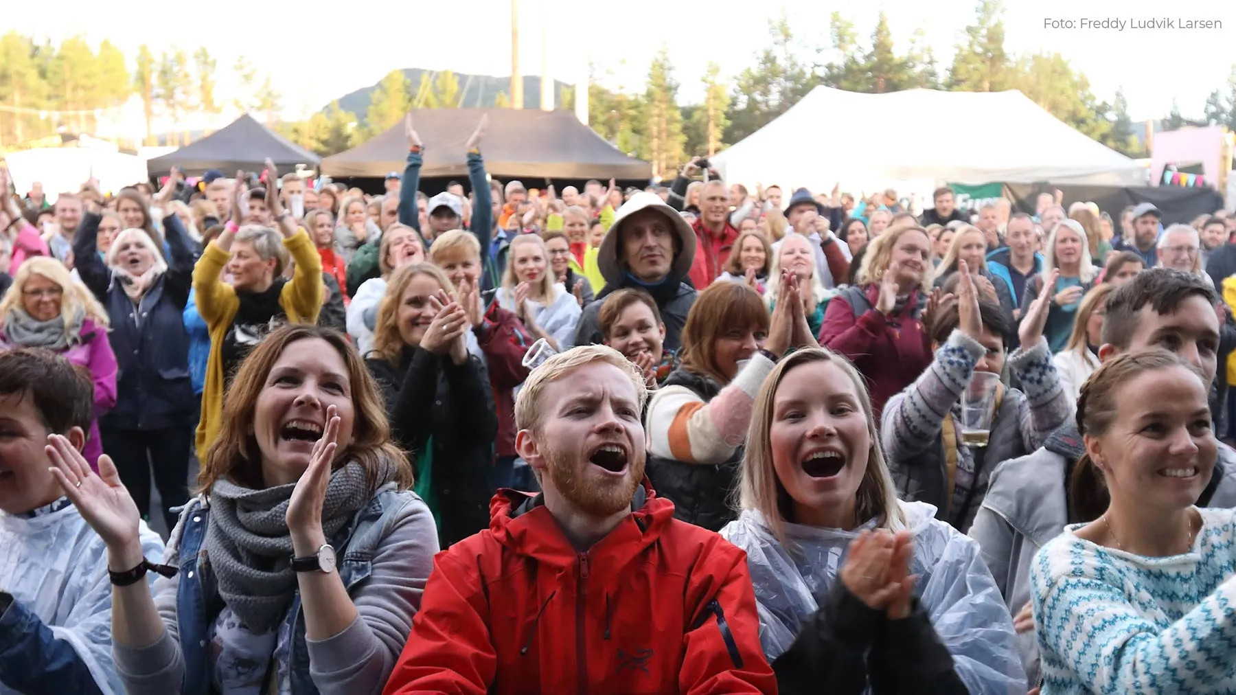 Glad folkemengde som klapper og jubler på en utendørs festival med telt i bakgrunnen.