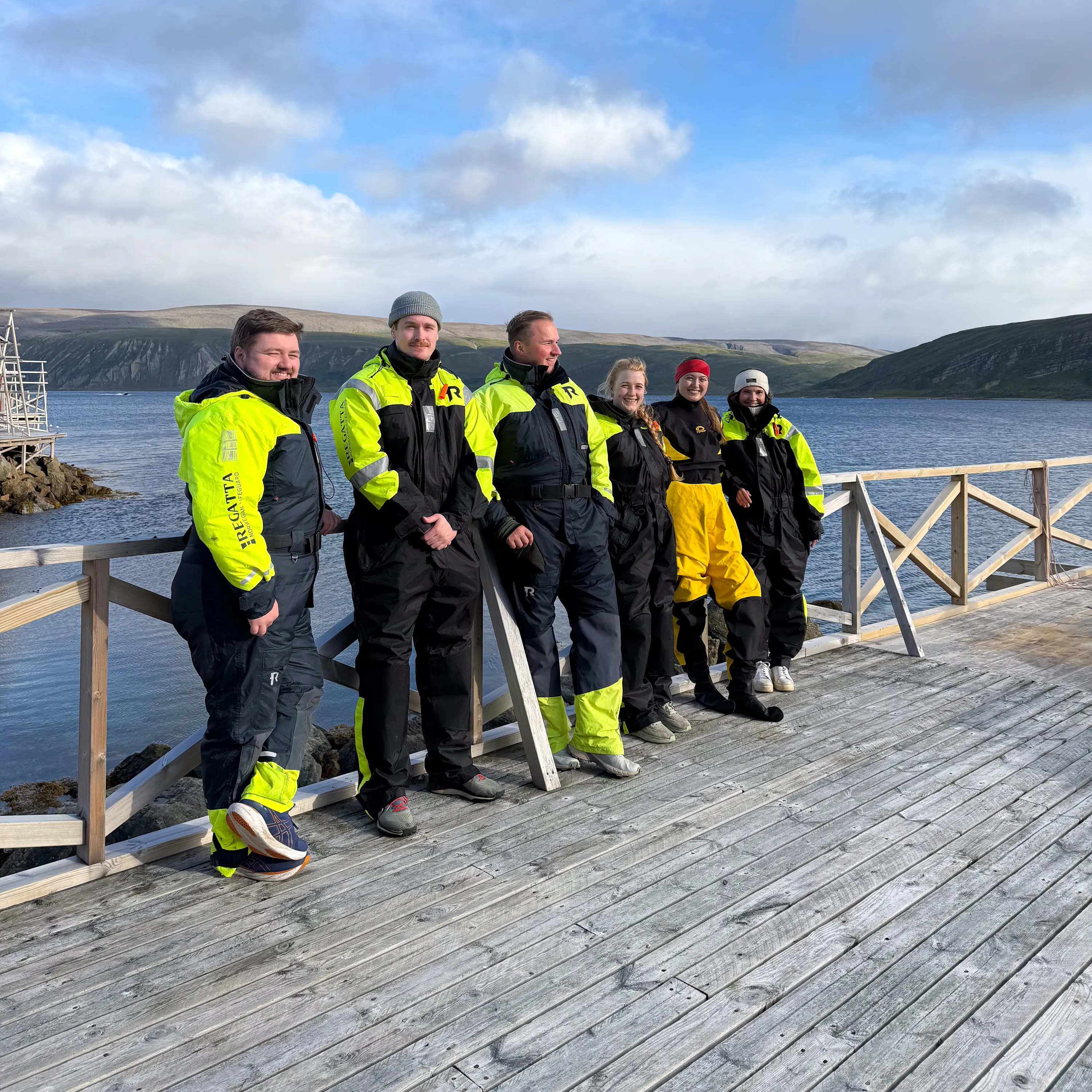 Seks personer i gule og svarte vanntette dresser står på en brygge med hav og fjell i bakgrunnen.