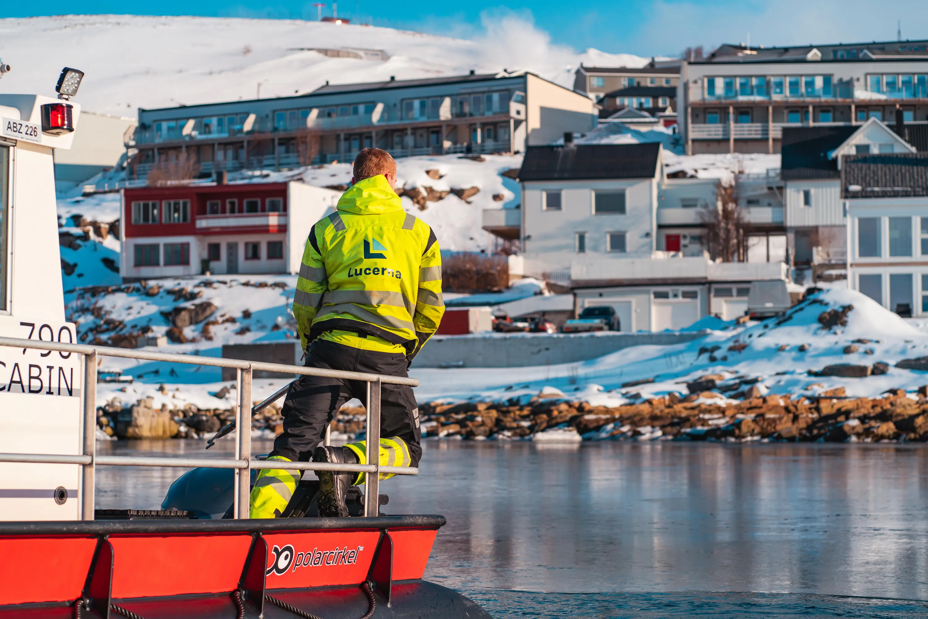 Person med gul refleksjakke på en båt ved iskaldt vann, med snødekte hus og fjell i bakgrunnen.