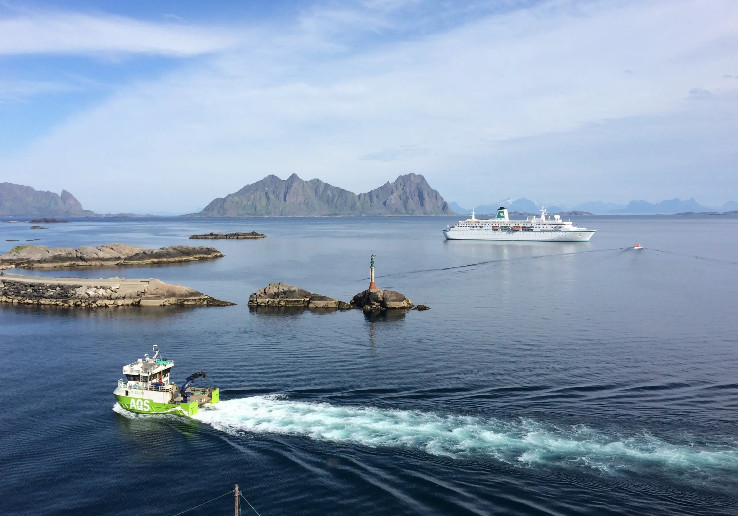 Grønt arbeidsfartøy og hvitt cruiseskip på rolig sjø med fjell i bakgrunnen under en blå himmel.