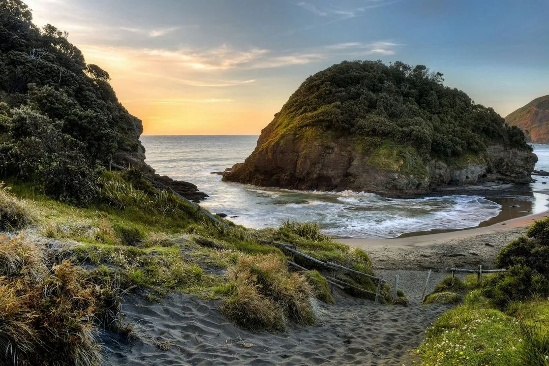 A sandy path leads to a serene beach scene with lush green hills, gentle waves, and a colorful sunset over the horizon.