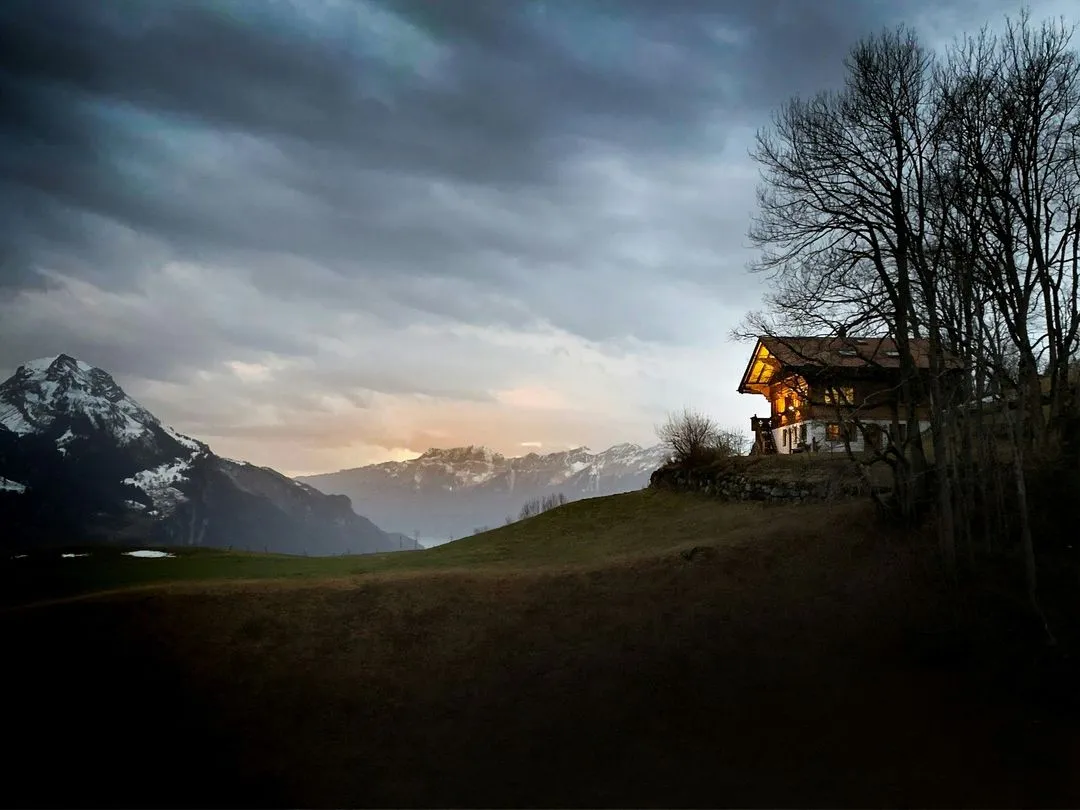 A cozy mountain house illuminated at dusk, surrounded by snow-capped peaks and dark, moody skies.