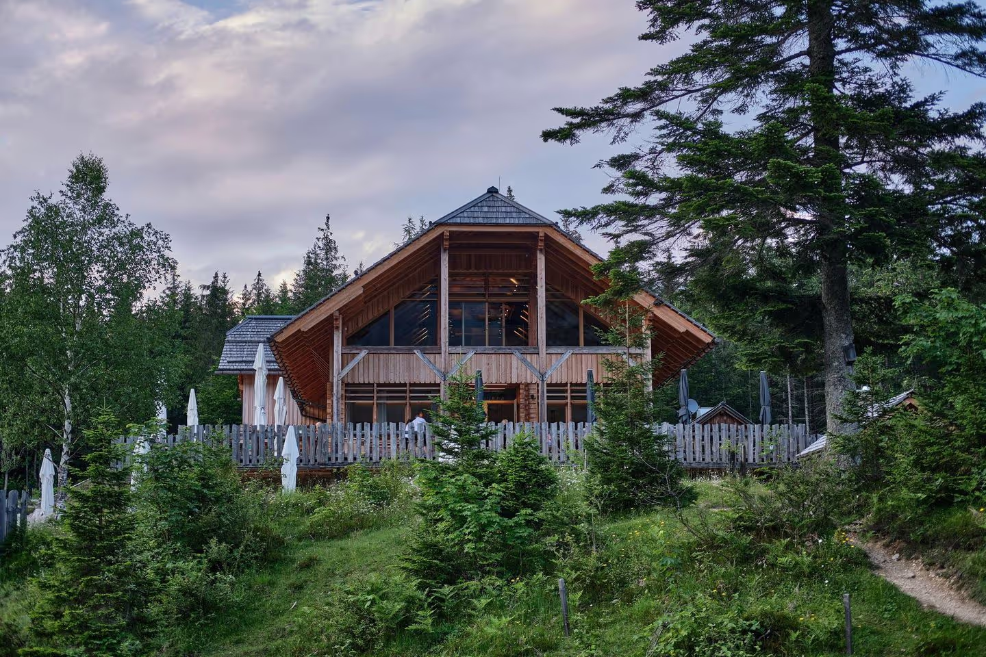 A modern wooden cabin with a large deck and glass facade, surrounded by lush greenery and trees under a cloudy sky.