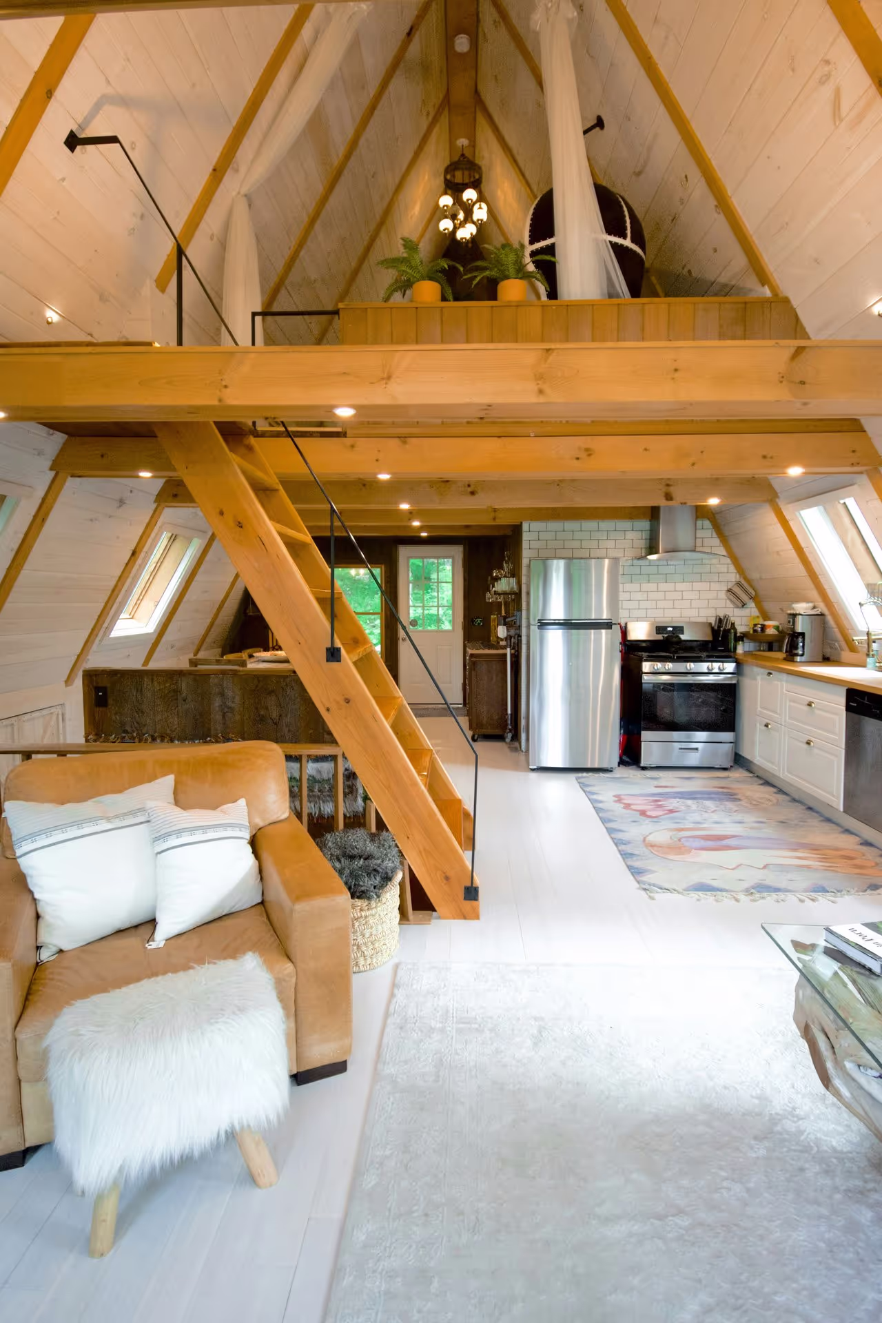 Cozy A-frame interior featuring a wooden staircase, leather armchair, and a modern kitchen with rustic decor and large windows.