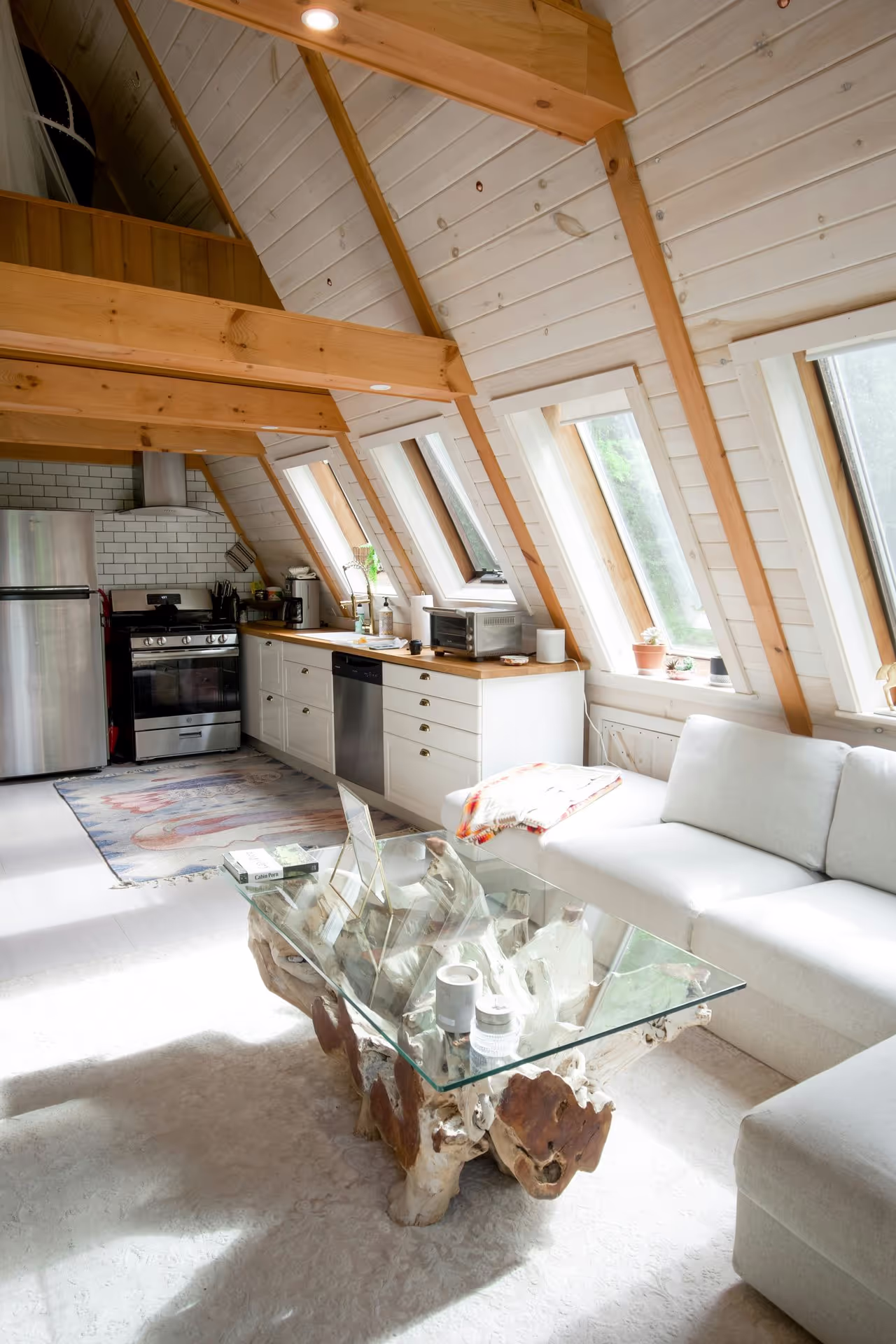 Brightly lit chalet interior featuring wooden beams, a white kitchen, and a glass-topped table on a plush rug, alongside a cozy sofa.