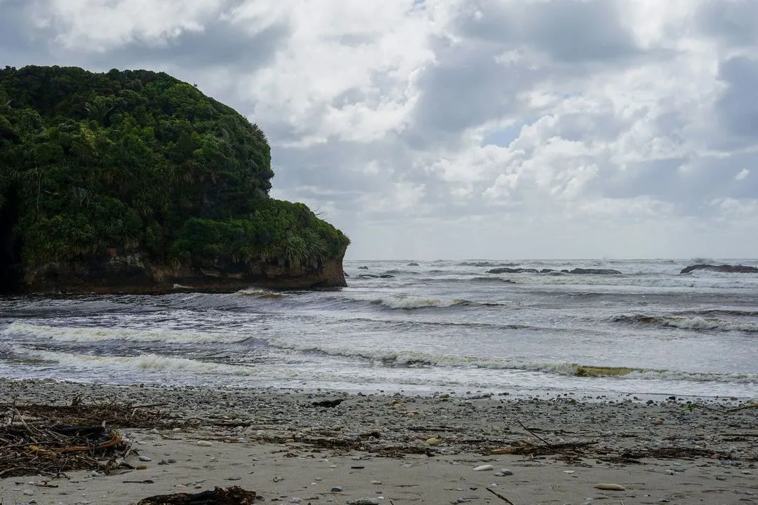A rocky coastline meets a turbulent sea under a cloudy sky, with waves gently lapping the sandy shore.