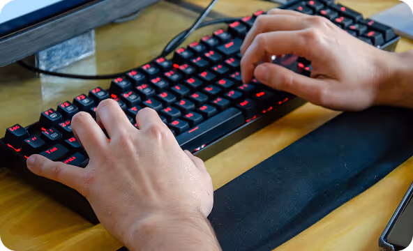 Hands typing on a black keyboard with red backlit keys on a wooden desk.