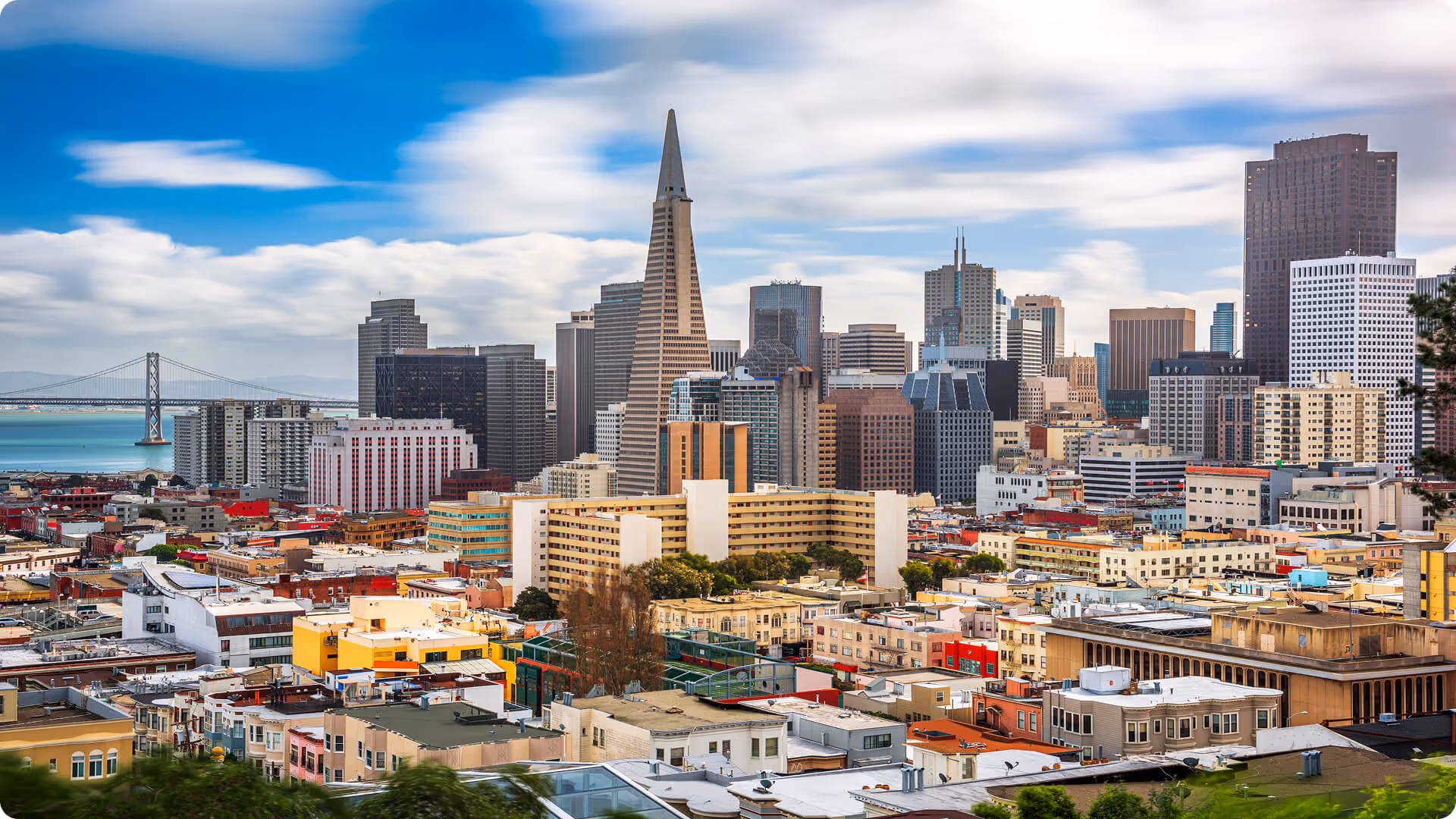 Panoramic view of San Francisco skyline featuring the Transamerica Pyramid and the Bay Bridge under a partly cloudy sky.