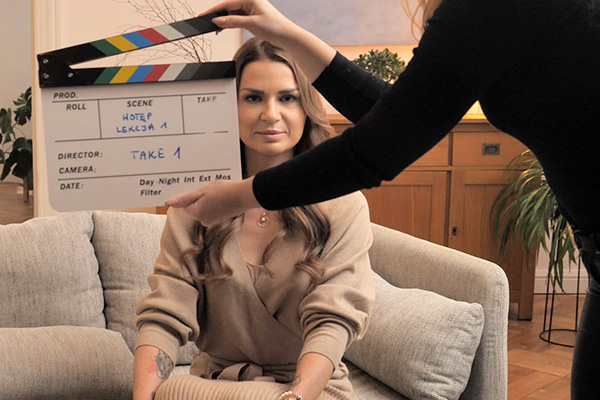 Woman sitting on a gray couch preparing for a film scene while a crew member holds a clapperboard in front of her.