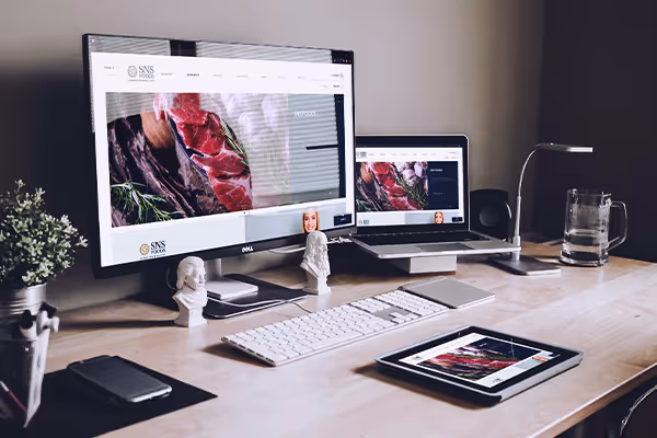 Workspace with a desktop monitor, laptop, and tablet all displaying the same image of raw meat with herbs on a wooden surface.