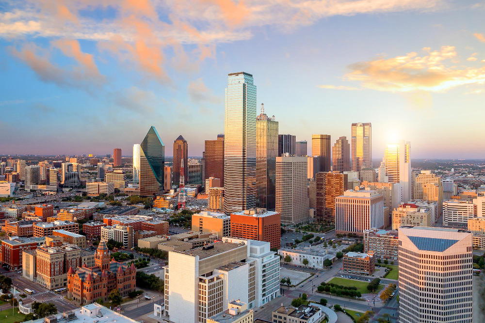 Aerial view of downtown Dallas skyline with modern skyscrapers illuminated by warm sunset light and soft clouds in the sky.