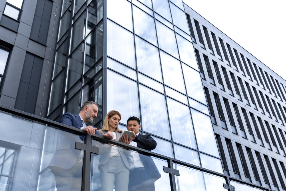 Three business professionals standing on a glass balcony outside a modern office building, looking at a tablet together.