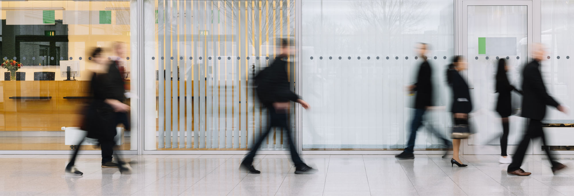 Blurred motion of people walking past a modern office entrance with glass doors and vertical blinds.