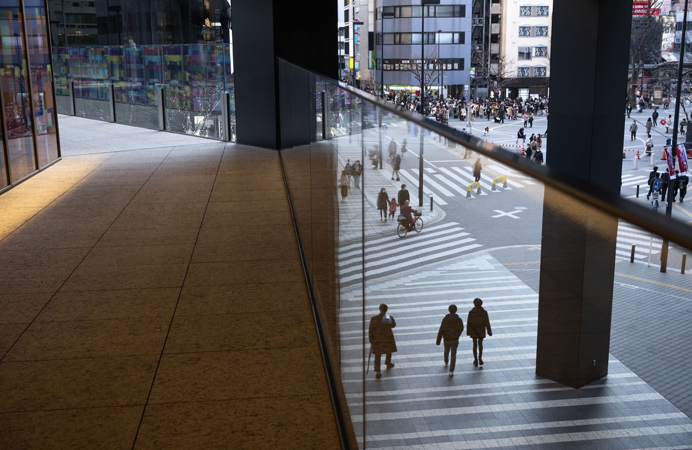 Urban crosswalk with pedestrians and a cyclist reflected in a glass wall alongside a tiled walkway.