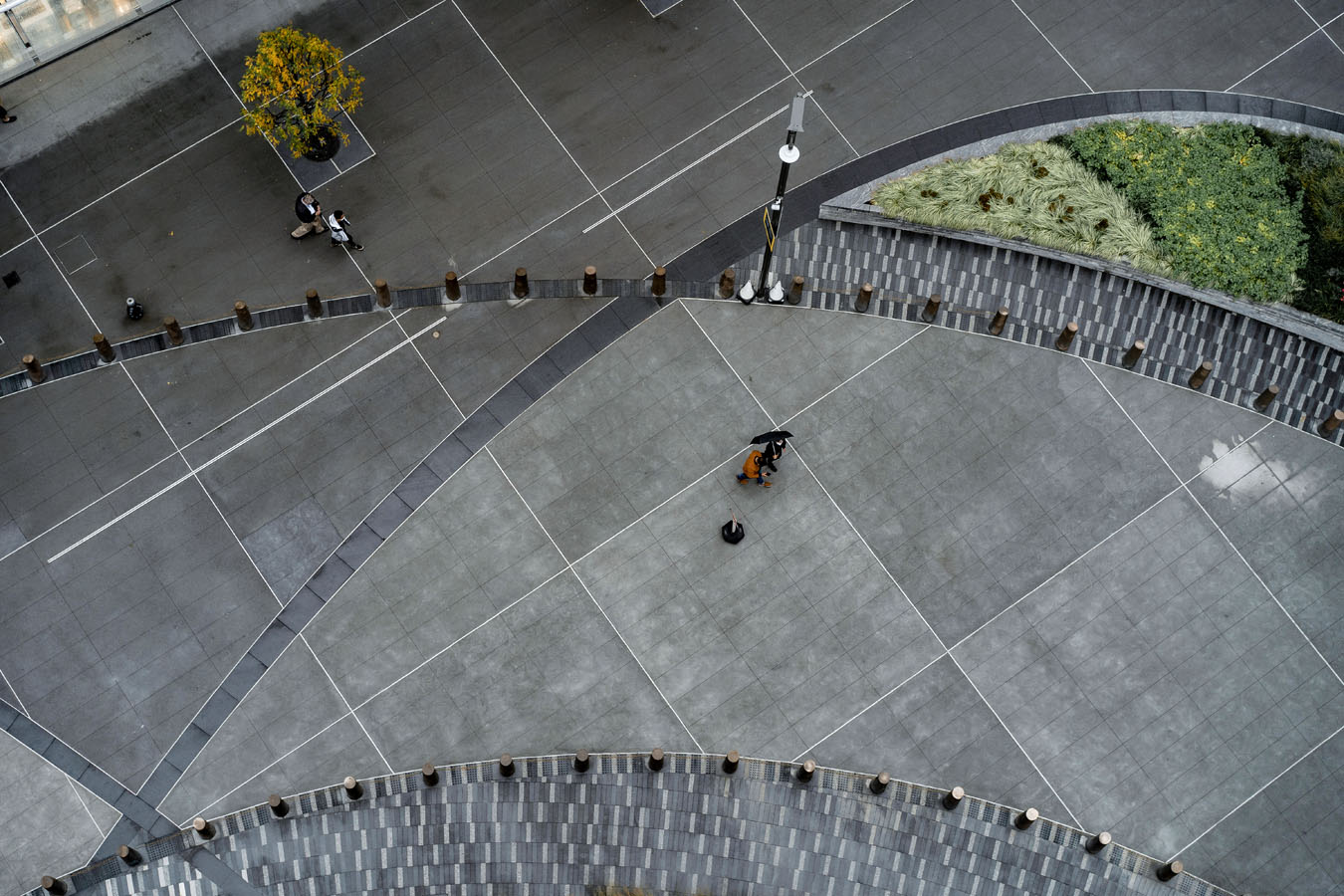 Aerial view of a paved urban plaza with two people walking under an umbrella and others near a tree and greenery.