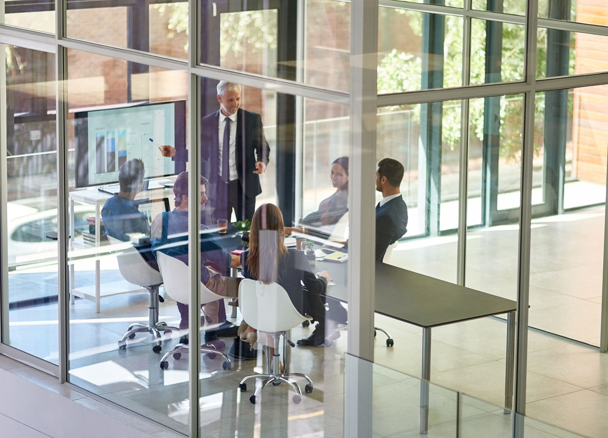 Businessman presenting data on a large screen to a group of colleagues in a modern glass-walled conference room.