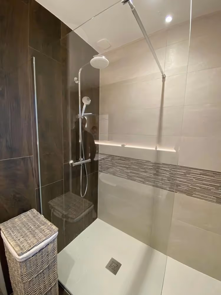 Modern walk-in shower with glass partition, rainfall and handheld showerheads, gray tiled walls, and a wicker laundry basket outside.