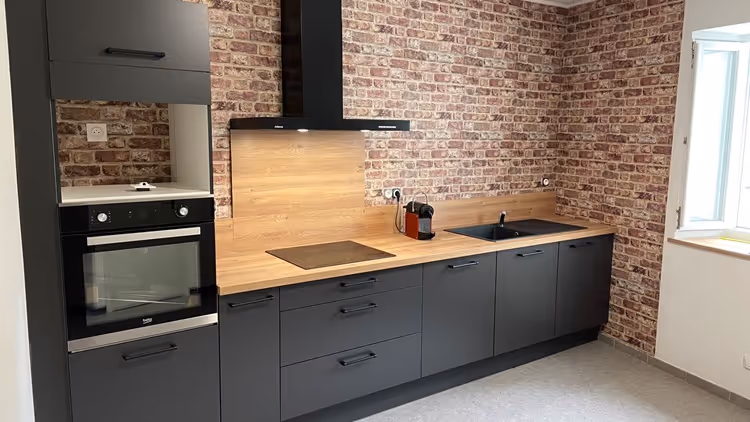 Modern kitchen with matte black cabinets, wooden countertop, built-in oven, electric cooktop, black sink, and brick-patterned walls.
