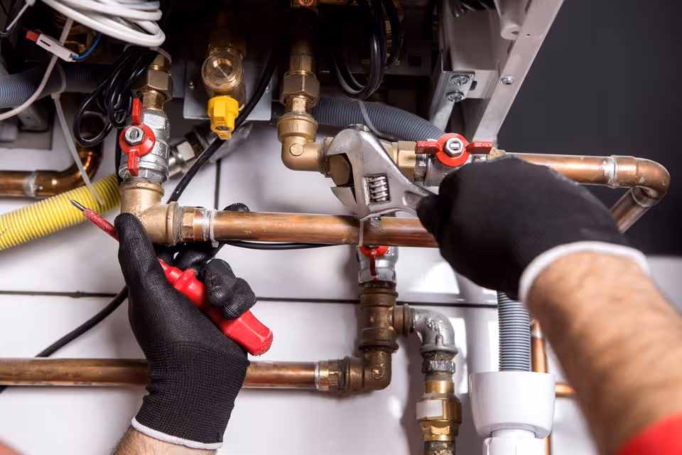 Gloved hands using a wrench and a screwdriver to repair copper plumbing pipes.