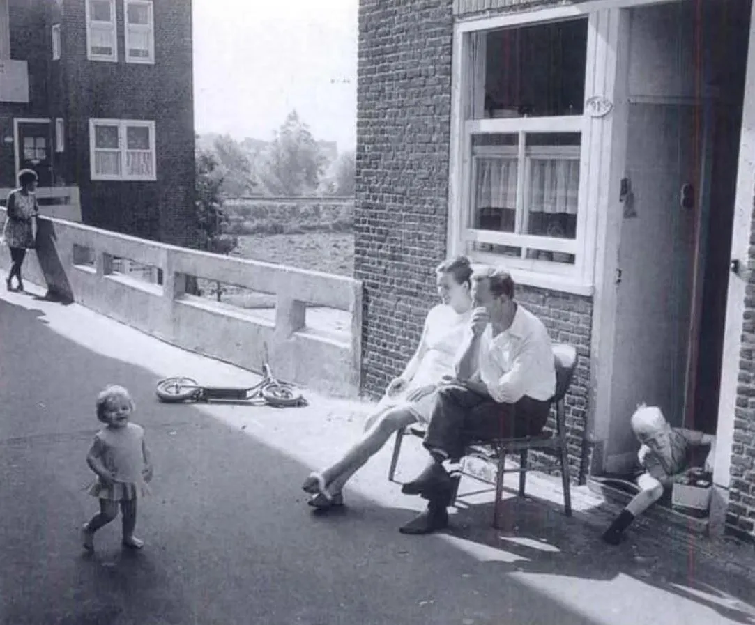 Črnobela fotografija prikazuje družino, ki sedi in se druži pred vhodom v delavsko stanovanje iz 60ih let, v prijetni senci - iz knjige Hermana Hertzbergerja