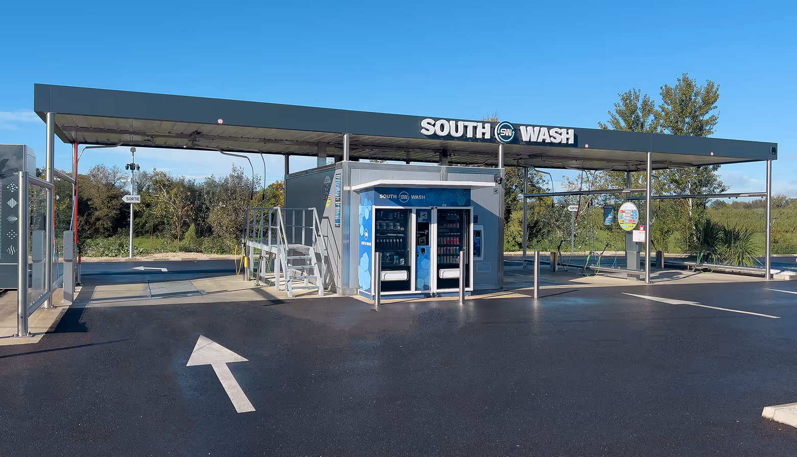Station de lavage automobile avec distributeurs automatiques sous un toit métallique et panneaux indiquant la sortie, sous ciel bleu.