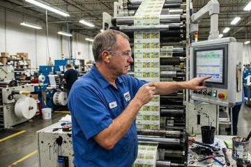 Man in a blue shirt operating a printing machine with color labels in an industrial setting.