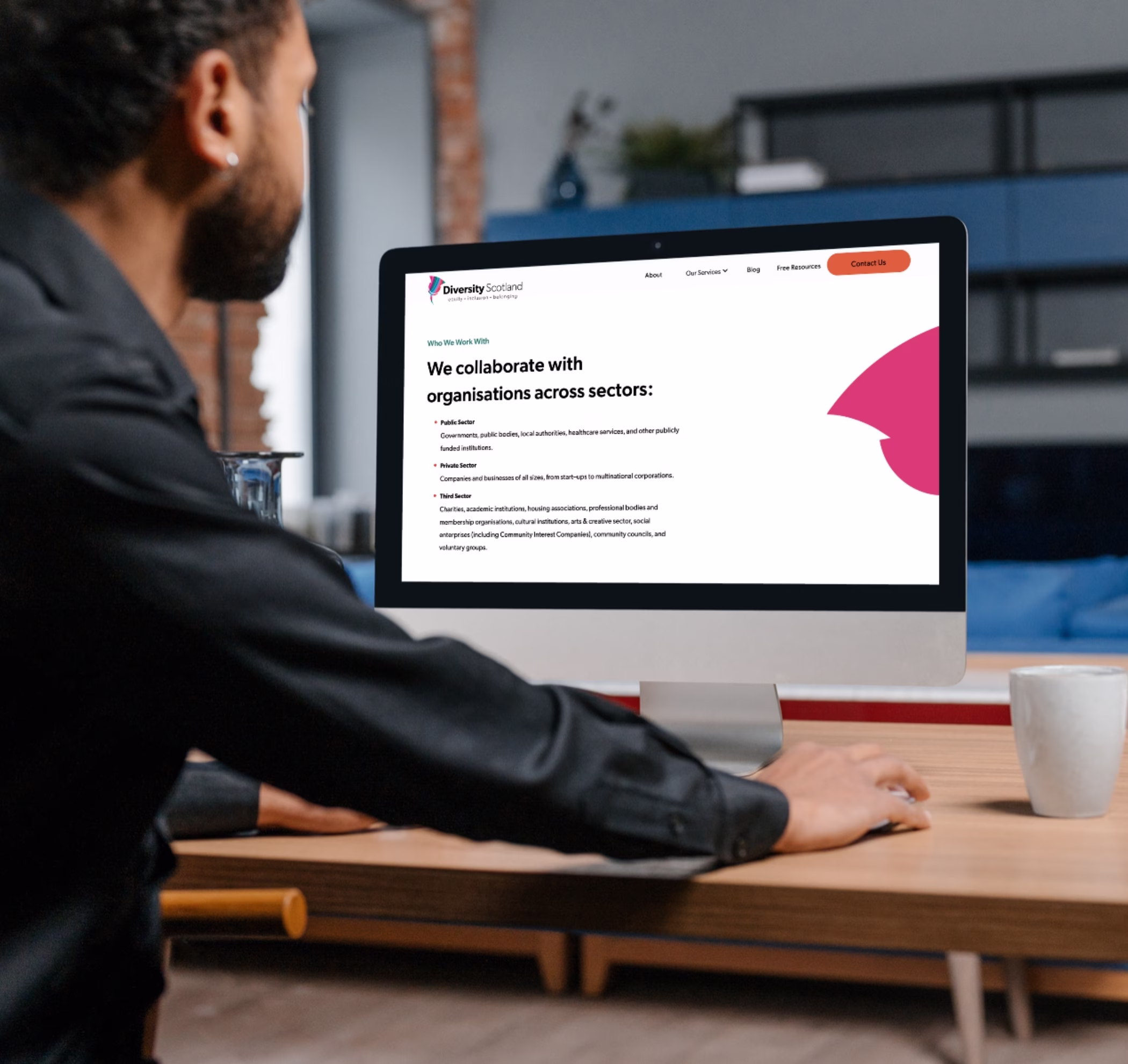 Person sitting at a desktop displaying the Diversity Scotland website, on a section titled "We collaborate with organisations across sectors" in clear dark text. The Diversity Scotland thistle icon in pink peeks out on the right of the screen.