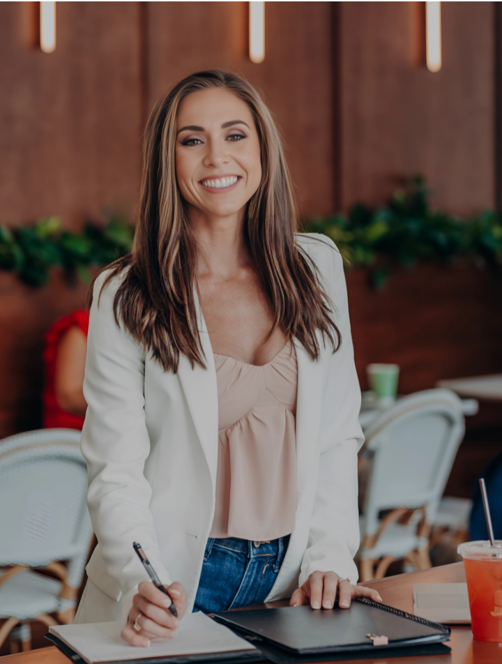 Smiling woman with long hair wearing a white blazer and light pink top, writing in a notebook at a table.