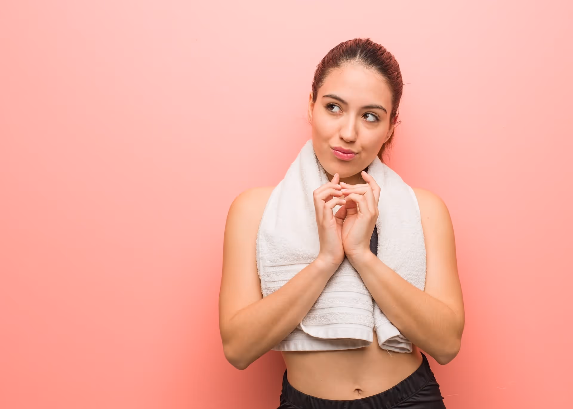 Young woman with a white towel around her neck standing against a pink background looking to the side thoughtfully.
