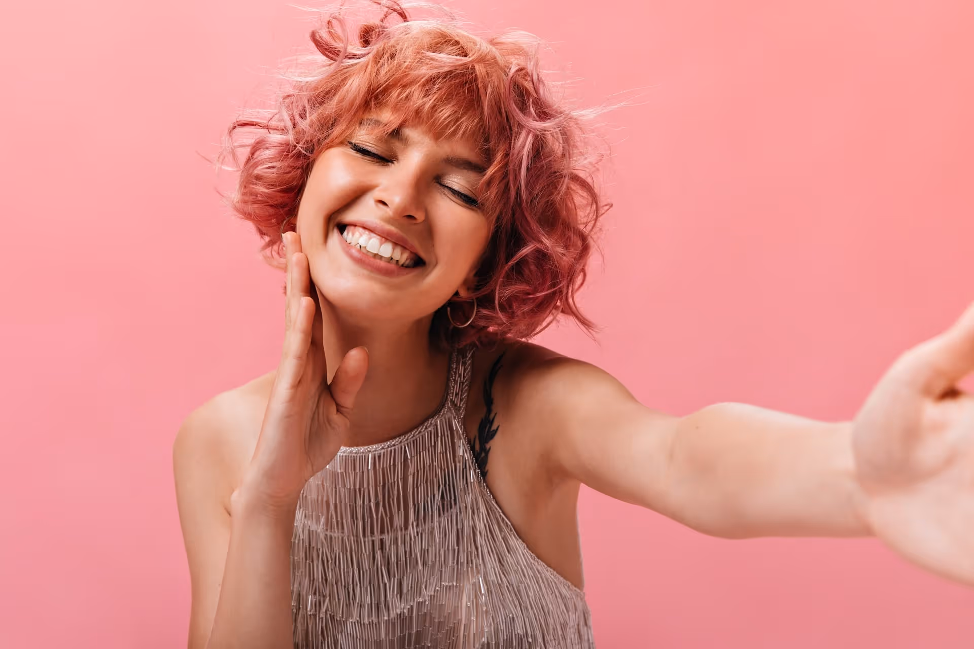Smiling woman with curly pink hair wearing a fringed top takes a selfie against a pink background.