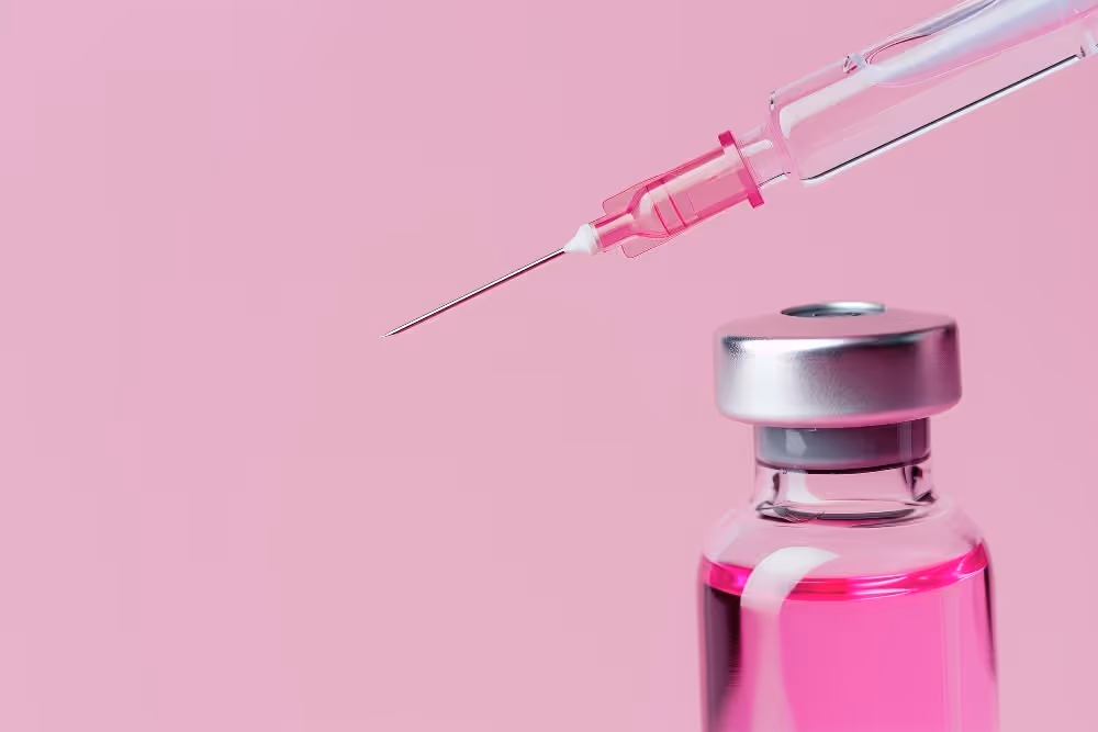 Syringe needle positioned above a vial filled with bright pink liquid against a pink background.