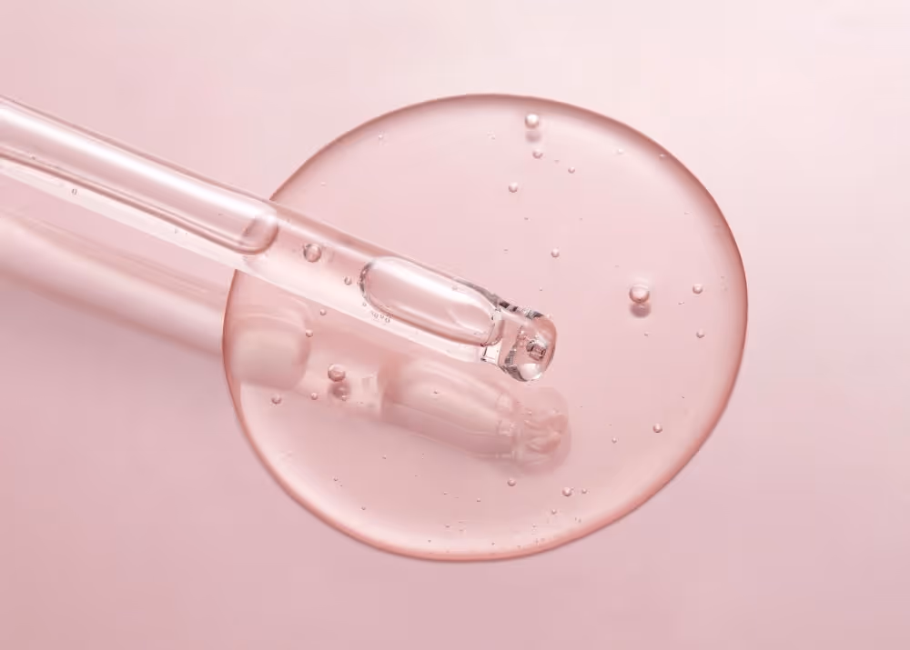 Close-up of a transparent dropper releasing a clear liquid drop on a pale pink surface.