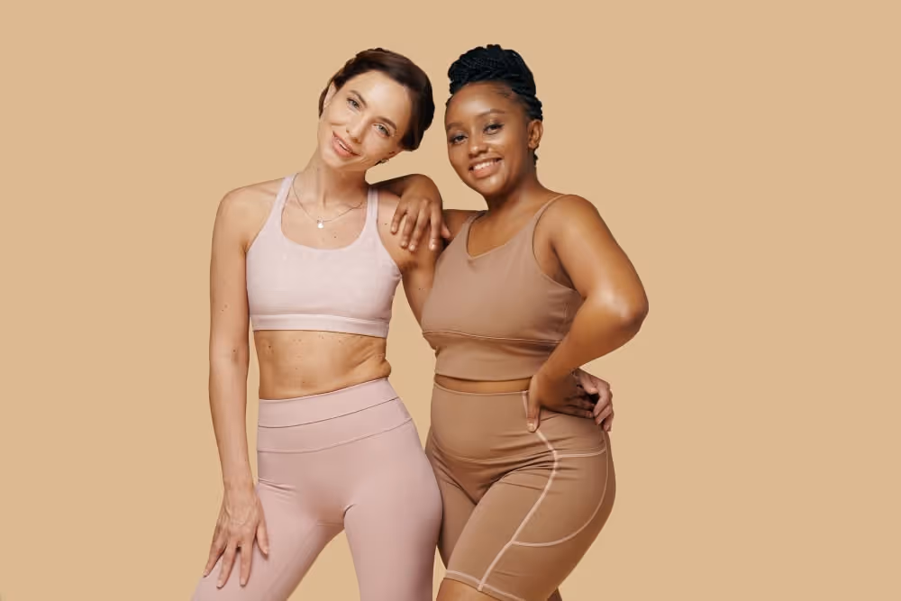 Two women in athletic wear posing together against a beige background, one with arm around the other's shoulder.